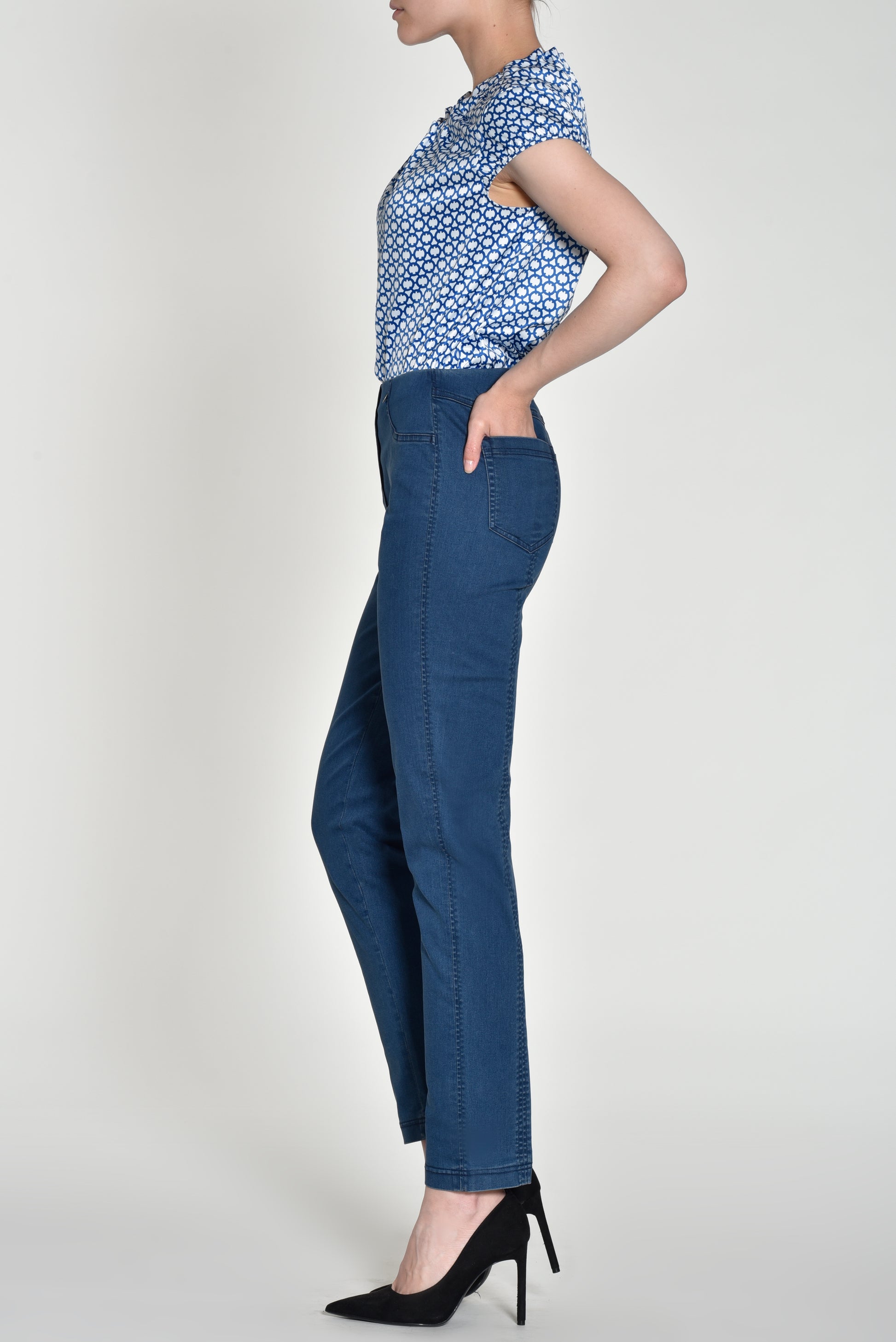 A woman shown in side profile wearing blue high-waisted straight-leg jeans, a blue and white patterned top, and black high heels against a light gray studio background.