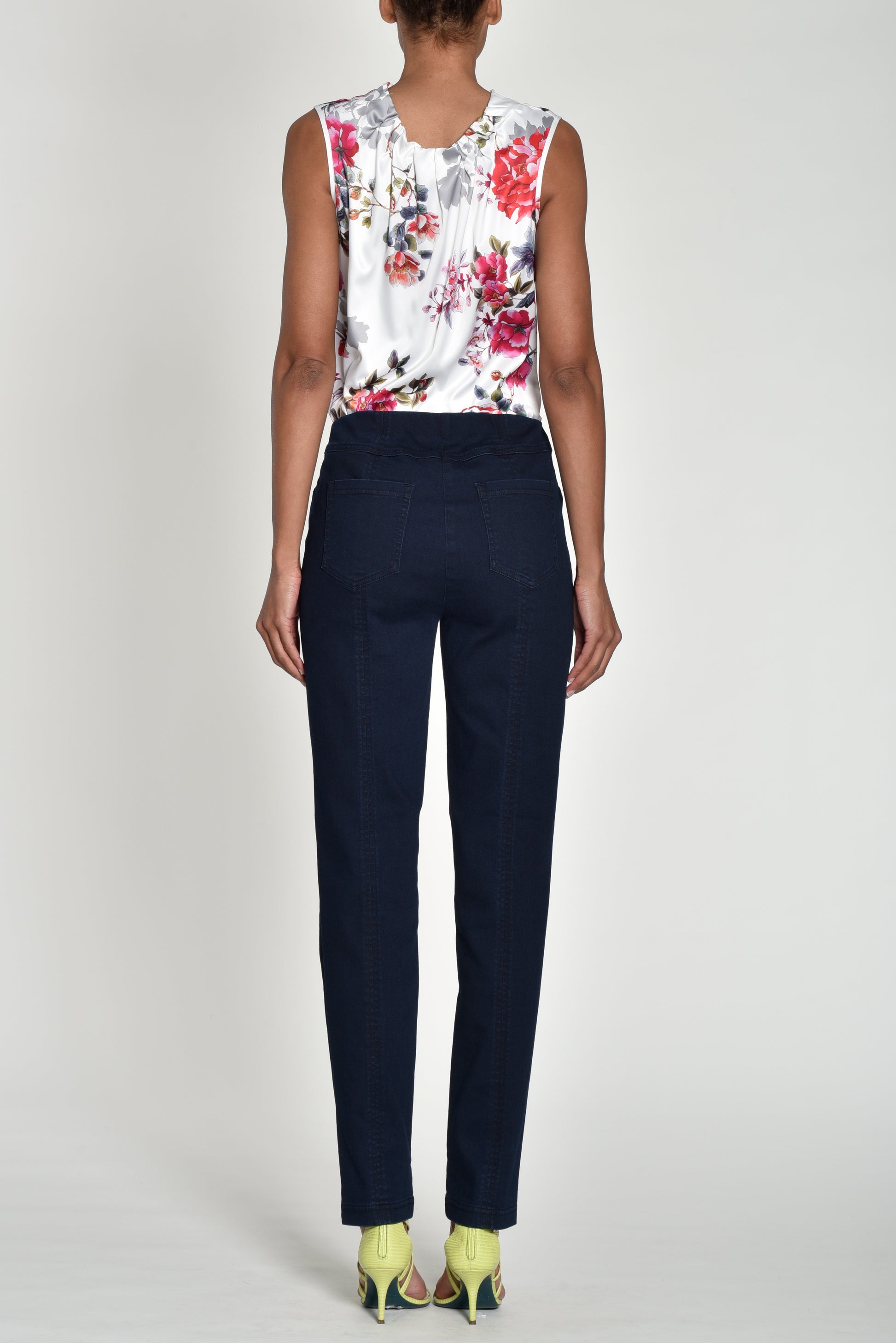 A model shown from the back wearing a sleeveless white top with a red and pink floral print, paired with dark navy blue trousers and lime green heels, against a light gray studio background.