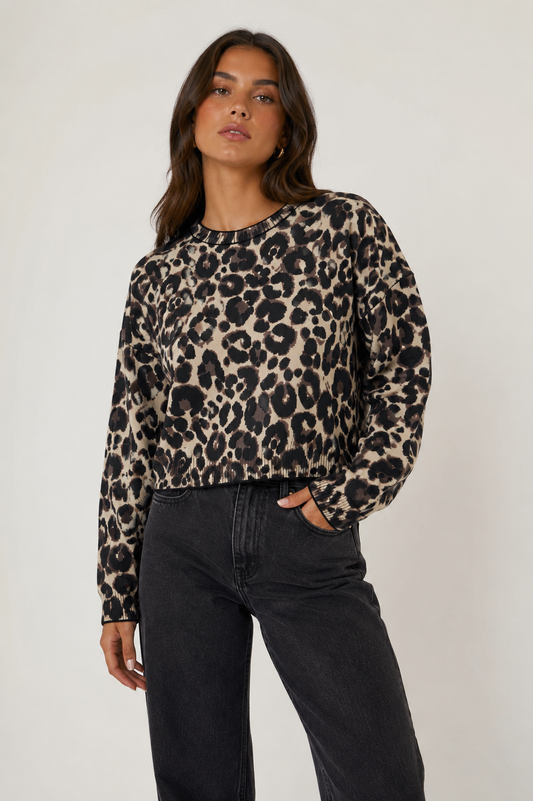 A woman modeling a long-sleeve crewneck top with a beige, brown, and black leopard print pattern, paired with dark charcoal gray jeans, against a plain light gray studio background.