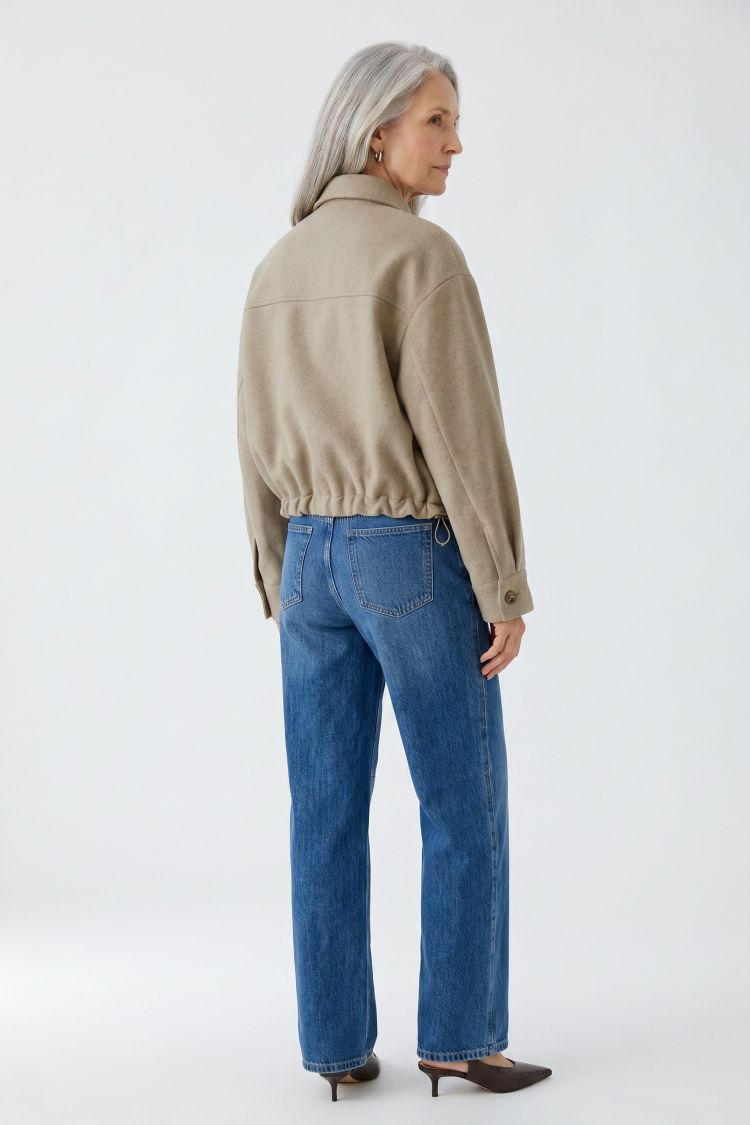 An older woman with silver hair is shown from the back wearing a beige cropped jacket and blue jeans, standing against a plain white studio background.