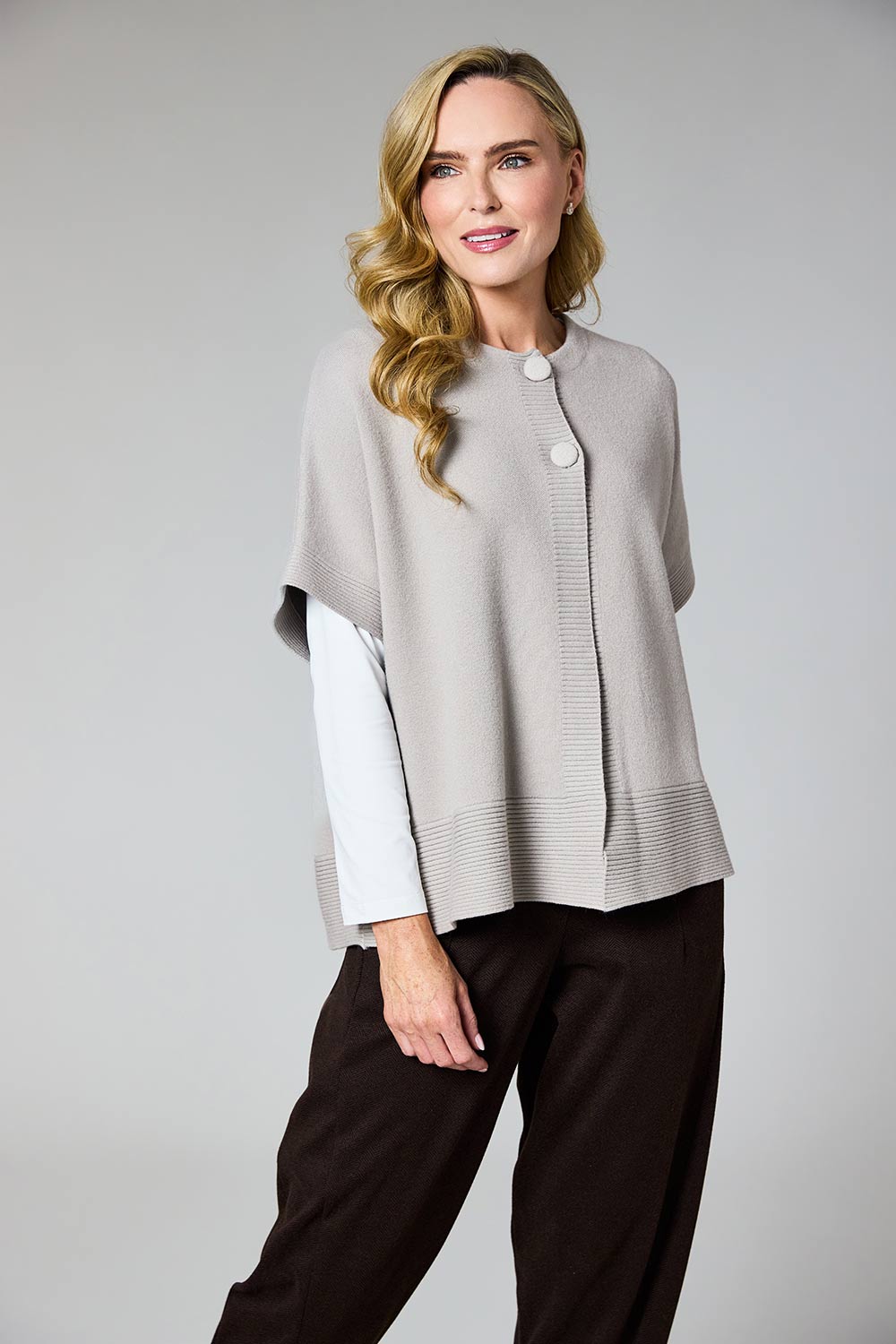 A woman modeling a light gray knit poncho-style cardigan with white buttons, layered over a white long-sleeve top and paired with dark brown pants, against a solid gray studio background.