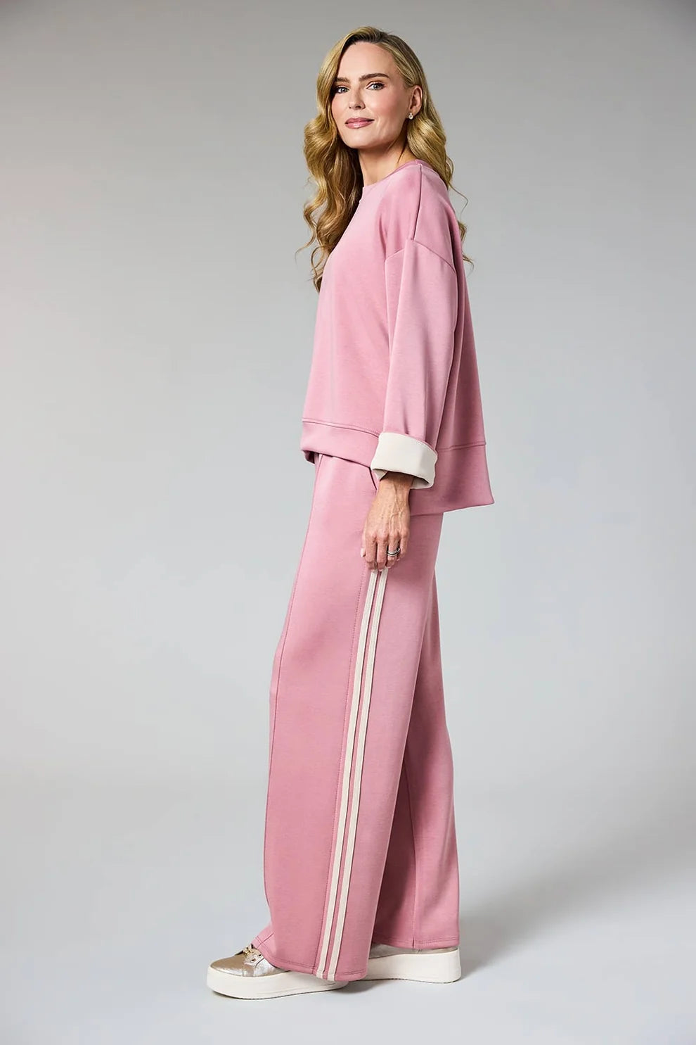 A woman modeling a matching pink lounge set consisting of a cropped sweatshirt with white ribbed cuffs and wide-leg pants with double white side stripes, photographed against a light gray studio background.