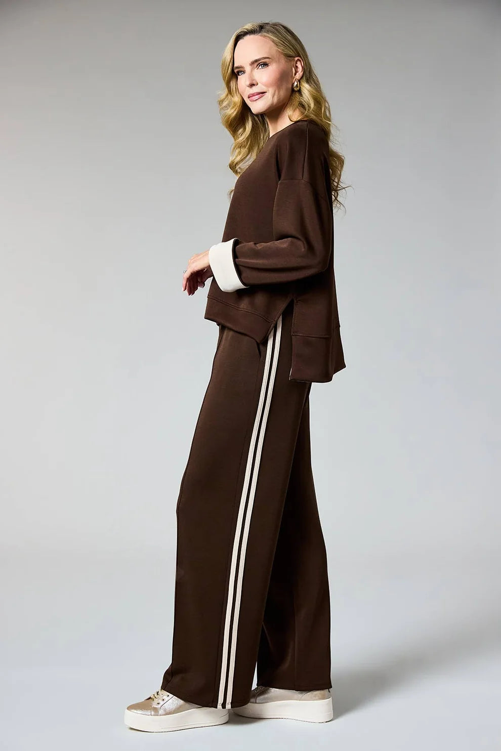 A woman modeling a brown two-piece lounge set consisting of a cropped long-sleeve top with contrasting white cuffs and matching wide-leg pants with double white side stripes, paired with white platform sneakers, against a light gray studio background.