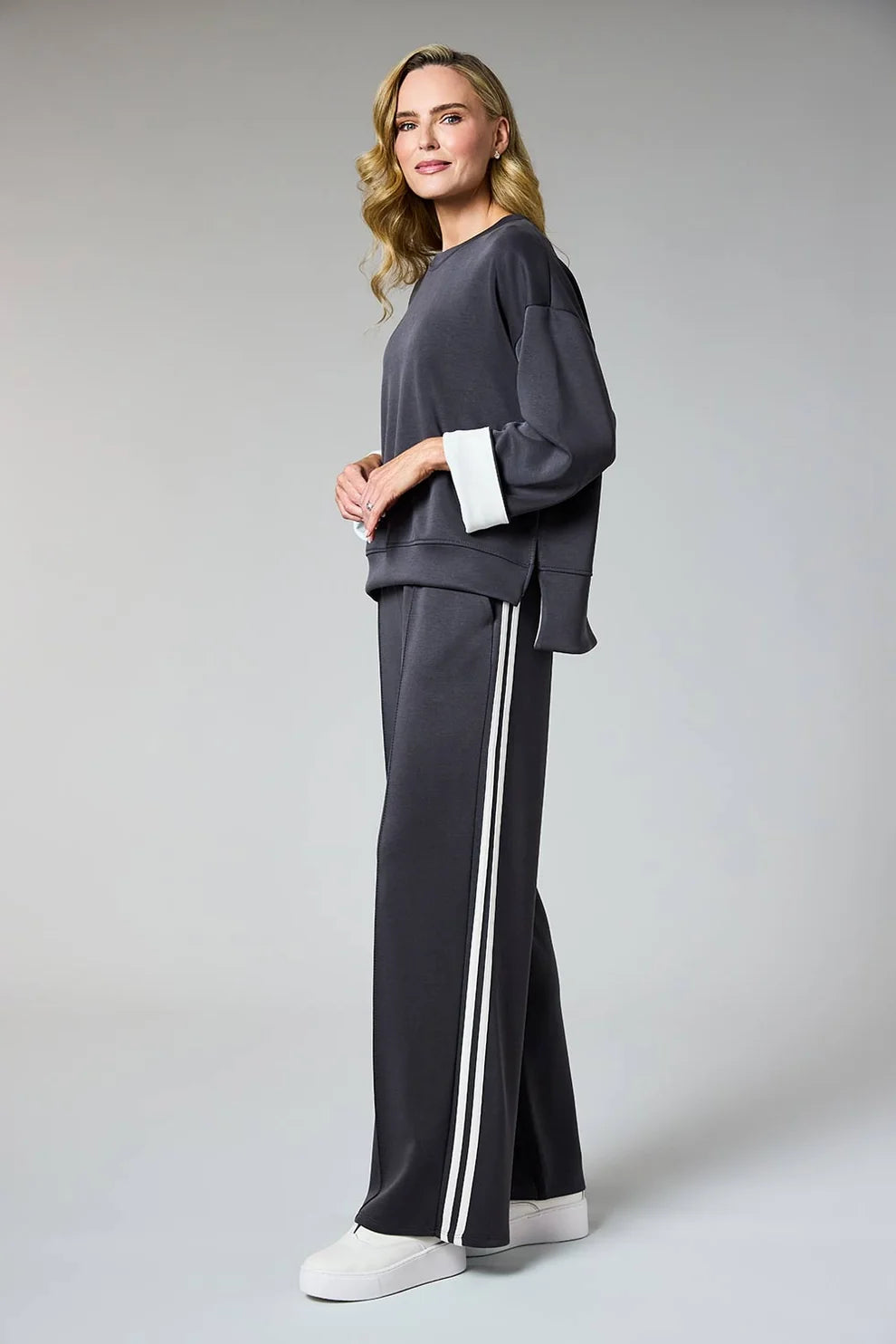 A woman modeling a dark gray two-piece lounge set consisting of a long-sleeve sweatshirt with contrasting white cuffs and matching wide-leg sweatpants with white side stripes, paired with white sneakers, against a light gray studio background.