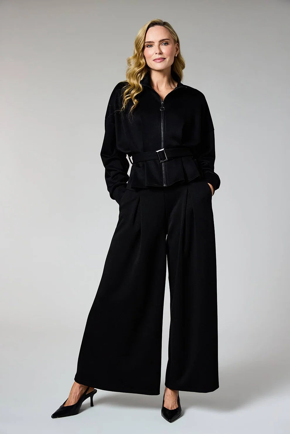 A woman modeling a black long-sleeve belted jacket with matching wide-leg trousers and black heels, photographed against a light gray studio background.