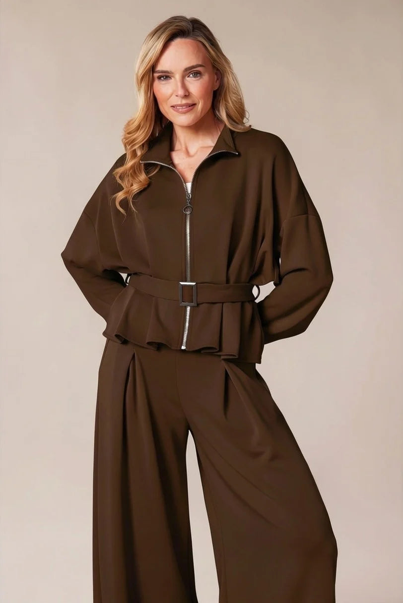 A woman modeling a dark brown two-piece jumpsuit with a zip-up long-sleeve top and matching wide-leg pants, set against a plain beige studio background.
