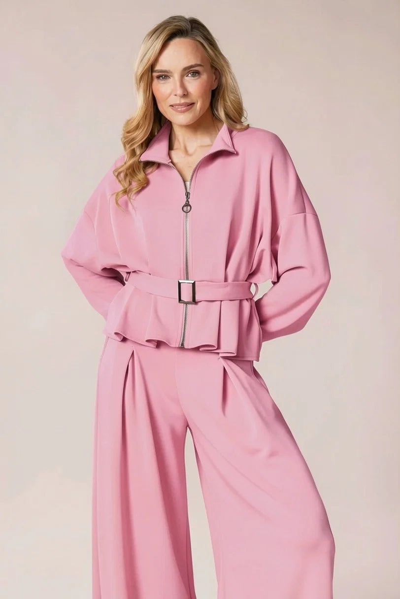 A woman modeling a pink two-piece jumpsuit set featuring a long-sleeve collared top with a front zipper and matching ruffled waist belt, paired with matching wide-leg trousers, against a solid light beige background.