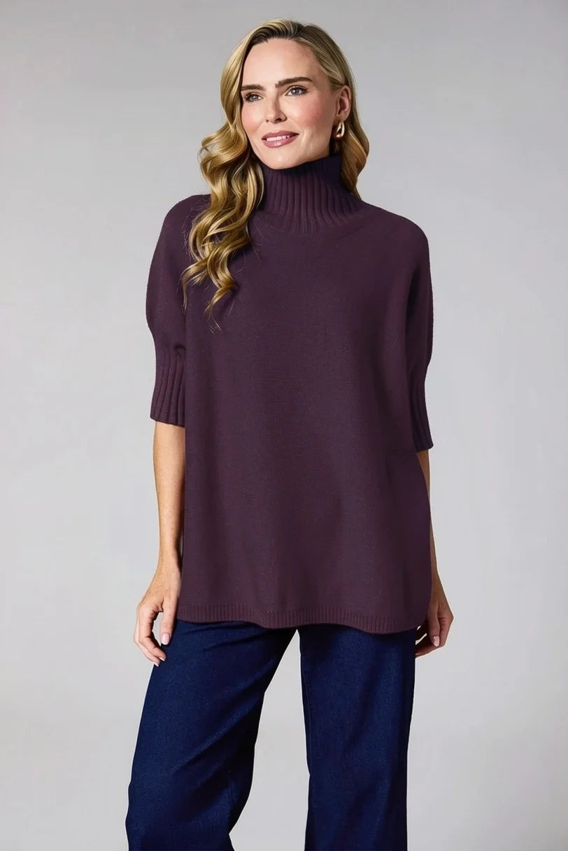 A woman modeling a mulberry-colored knit turtleneck sweater with elbow-length sleeves, paired with dark blue pants, against a solid light gray background.