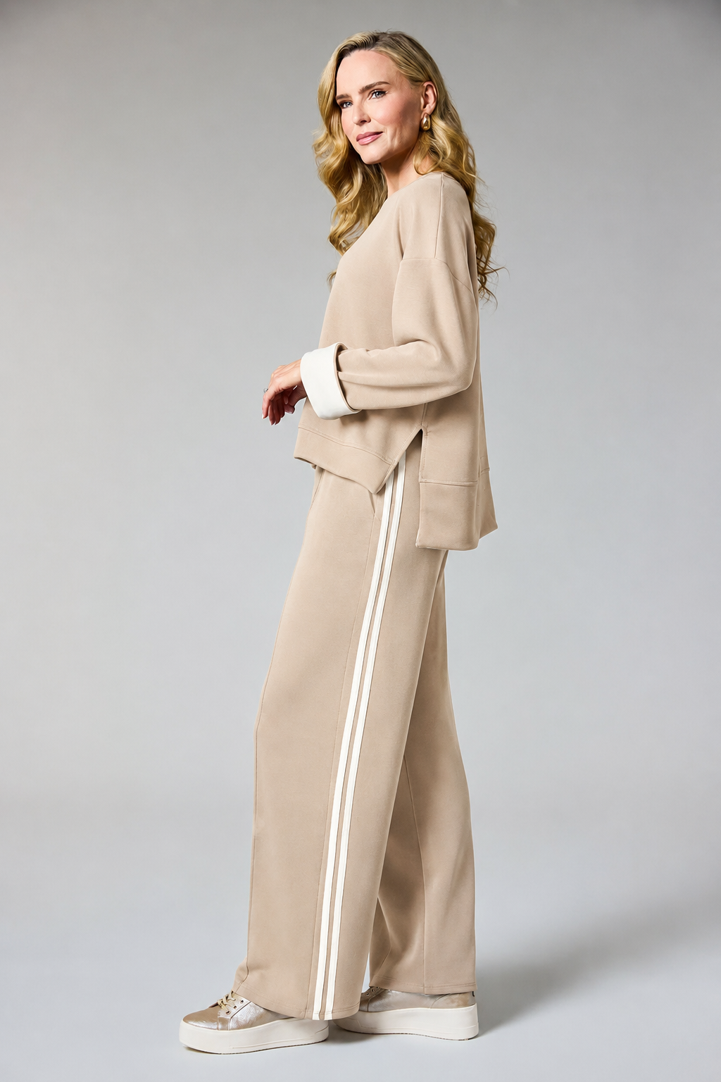 A woman modeling a beige two-piece lounge set consisting of a cropped long-sleeve top with white contrast cuffs and matching wide-leg pants with white side stripes, paired with white platform sneakers, against a light gray studio background.