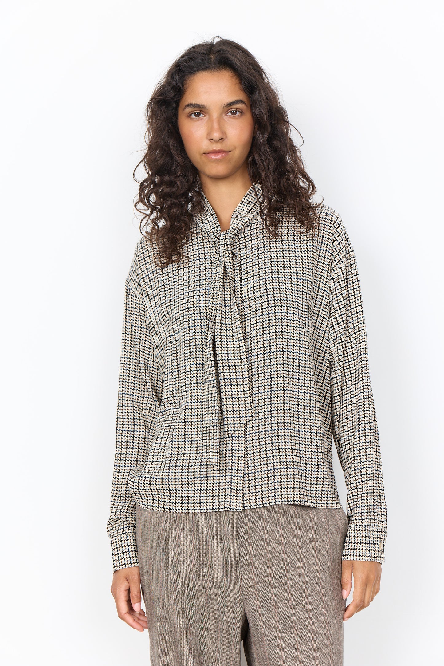 A woman with curly dark hair wearing a beige and black checkered long-sleeve blouse with a tie-neck detail, paired with gray trousers, standing against a plain white background.