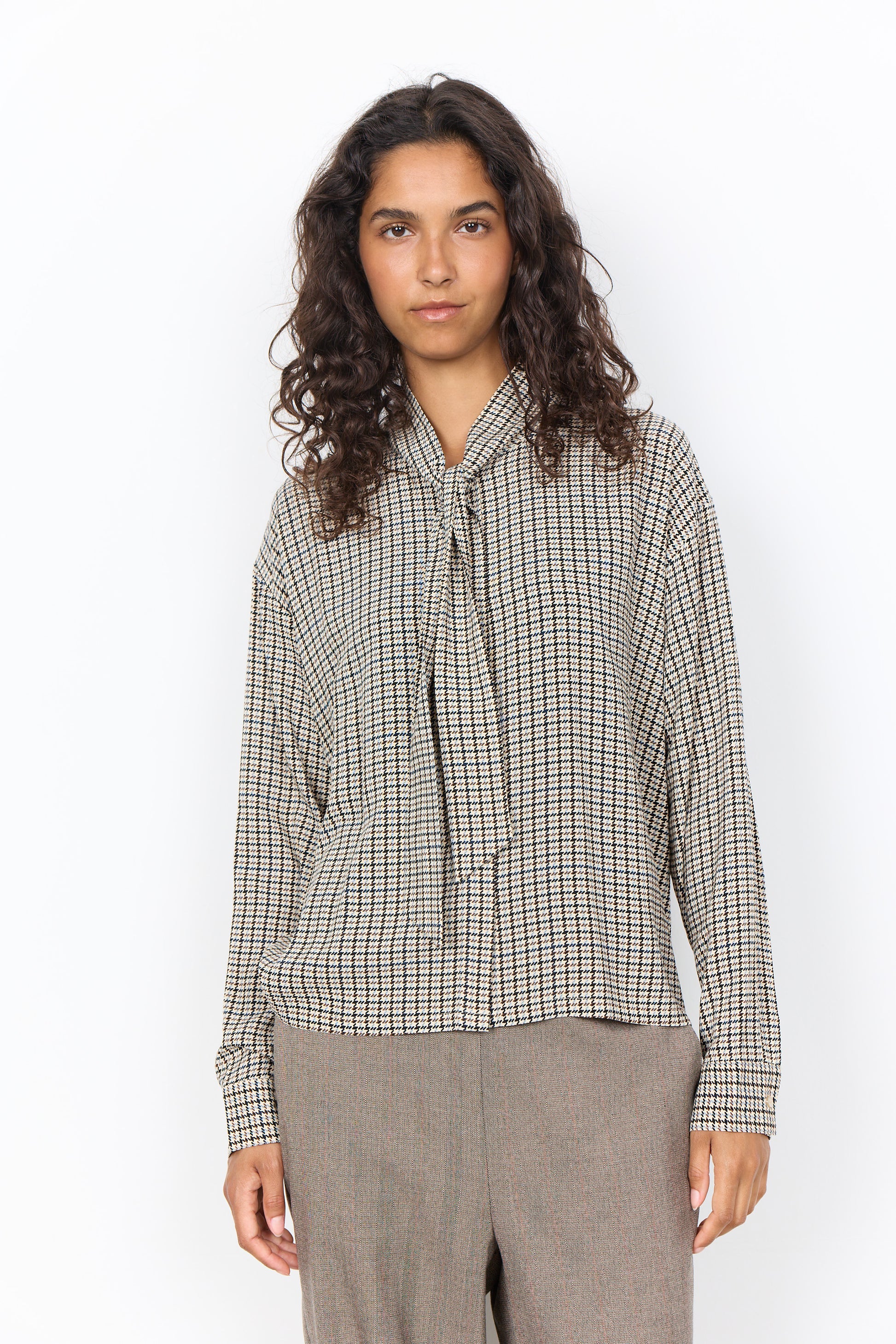 A woman with curly dark hair wearing a beige and black checkered long-sleeve blouse with a tie-neck detail, paired with gray trousers, standing against a plain white background.