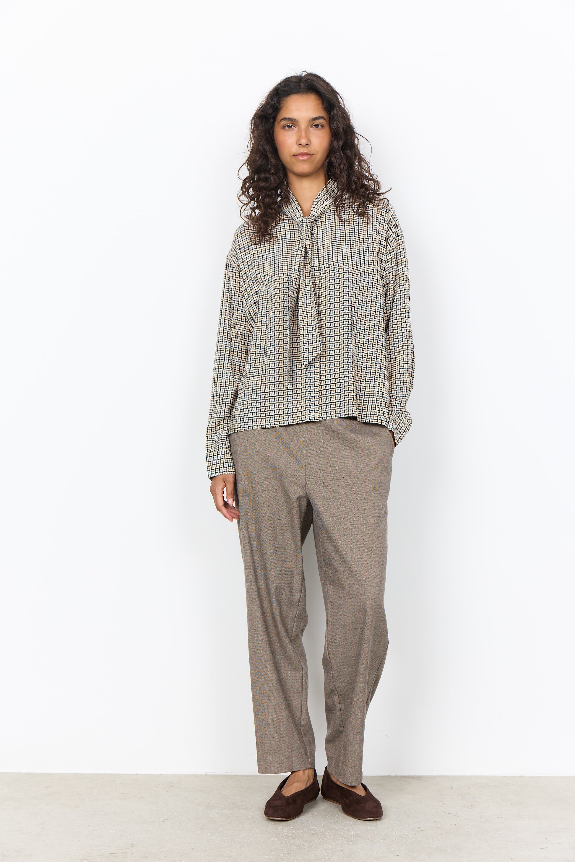 A model stands against a plain white wall wearing a long-sleeve button-up shirt with a small-scale check pattern in beige and brown, featuring a tie-neck detail, paired with loose-fit taupe trousers and dark brown loafers.