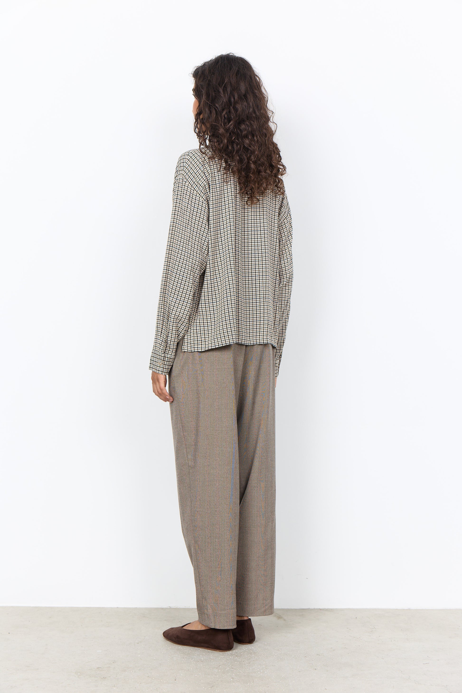 A woman stands with her back to the camera wearing a beige and brown gingham long-sleeve shirt and matching wide-leg trousers, paired with dark brown flat shoes, against a plain white wall.