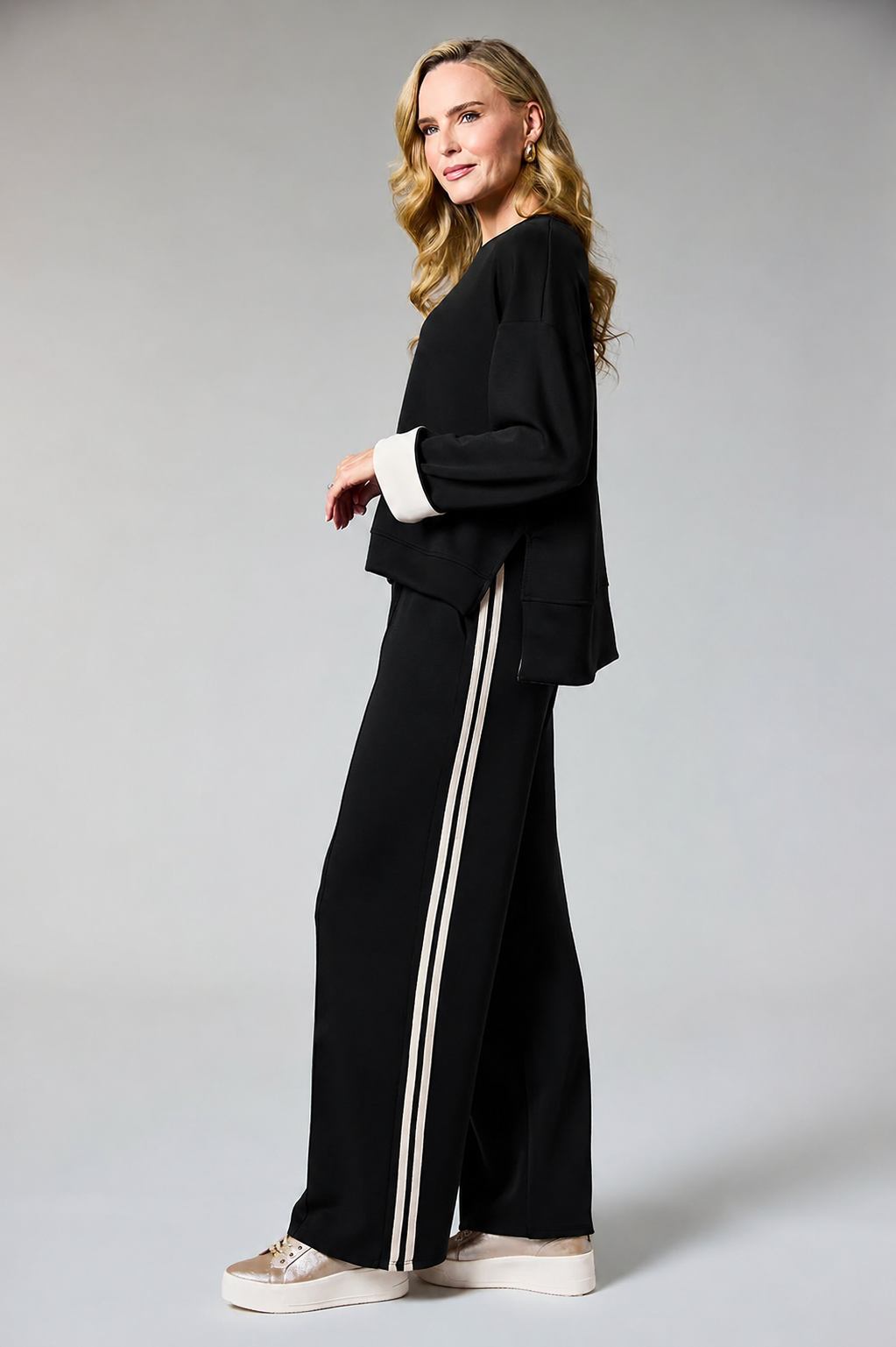 A woman modeling a black two-piece lounge set with white contrast trim, featuring a cropped long-sleeve top and matching wide-leg pants with double white side stripes, paired with beige platform sneakers against a light gray studio background.