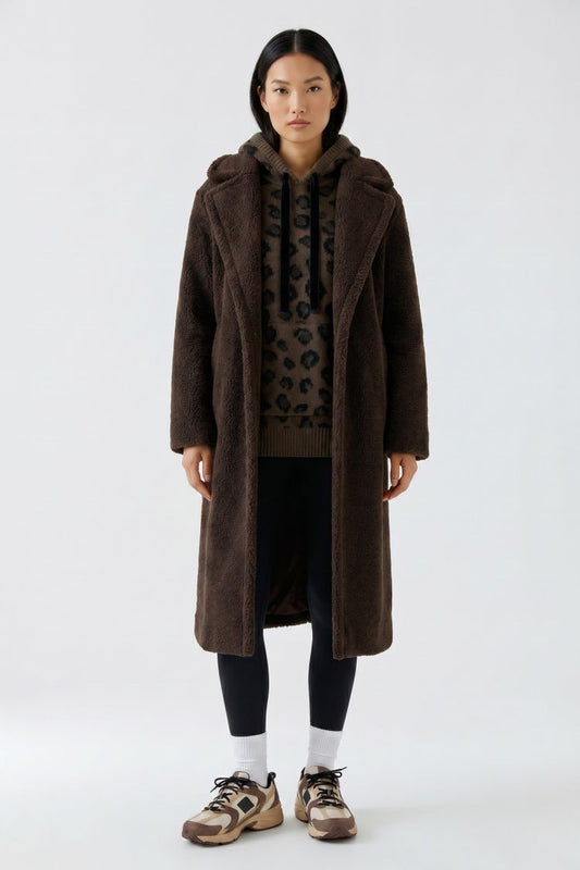 A model stands against a light gray studio background wearing a long dark brown teddy coat over a leopard-print top, black leggings, white socks, and brown sneakers.