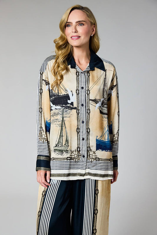 A woman modeling a long-sleeve button-front shirt with a beige and black nautical print featuring sailboats and chain motifs, paired with dark striped pants, against a neutral gray studio background.