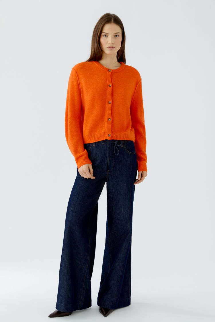 A model wearing a bright orange knit cardigan with buttons and dark blue wide-leg jeans, standing against a light gray studio background.