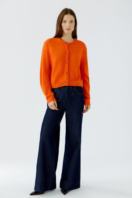 A model wearing a bright orange knit cardigan with buttons and dark blue wide-leg jeans, standing against a light gray studio background.