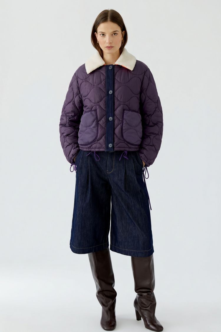 A model stands against a light gray studio background wearing a dark purple quilted puffer jacket with a cream-colored collar, dark blue wide-leg denim pants, and dark brown knee-high leather boots.
