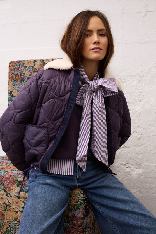 A woman models a dark purple quilted puffer jacket with a cream faux-fur collar, worn over a striped shirt and blue jeans, seated against a white brick wall.