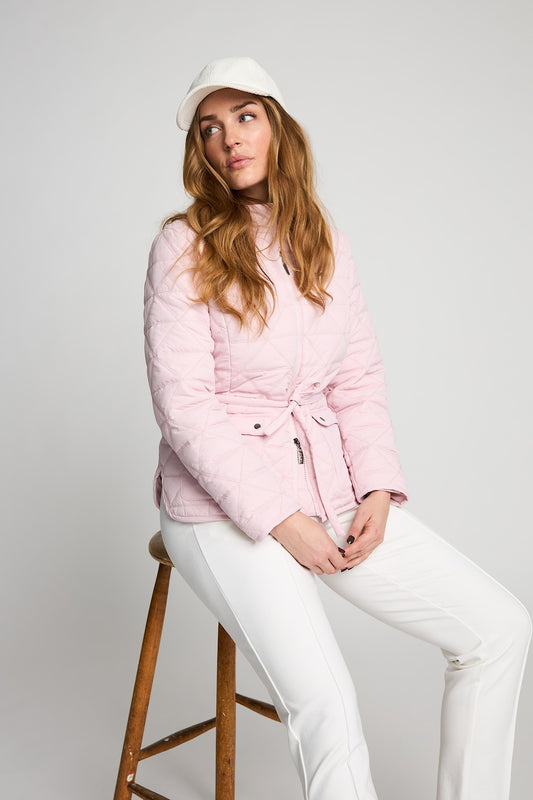 A woman wearing a light pink quilted puffer jacket with a tie waist, white pants, and a white cap, sitting on a wooden stool against a light gray studio background.