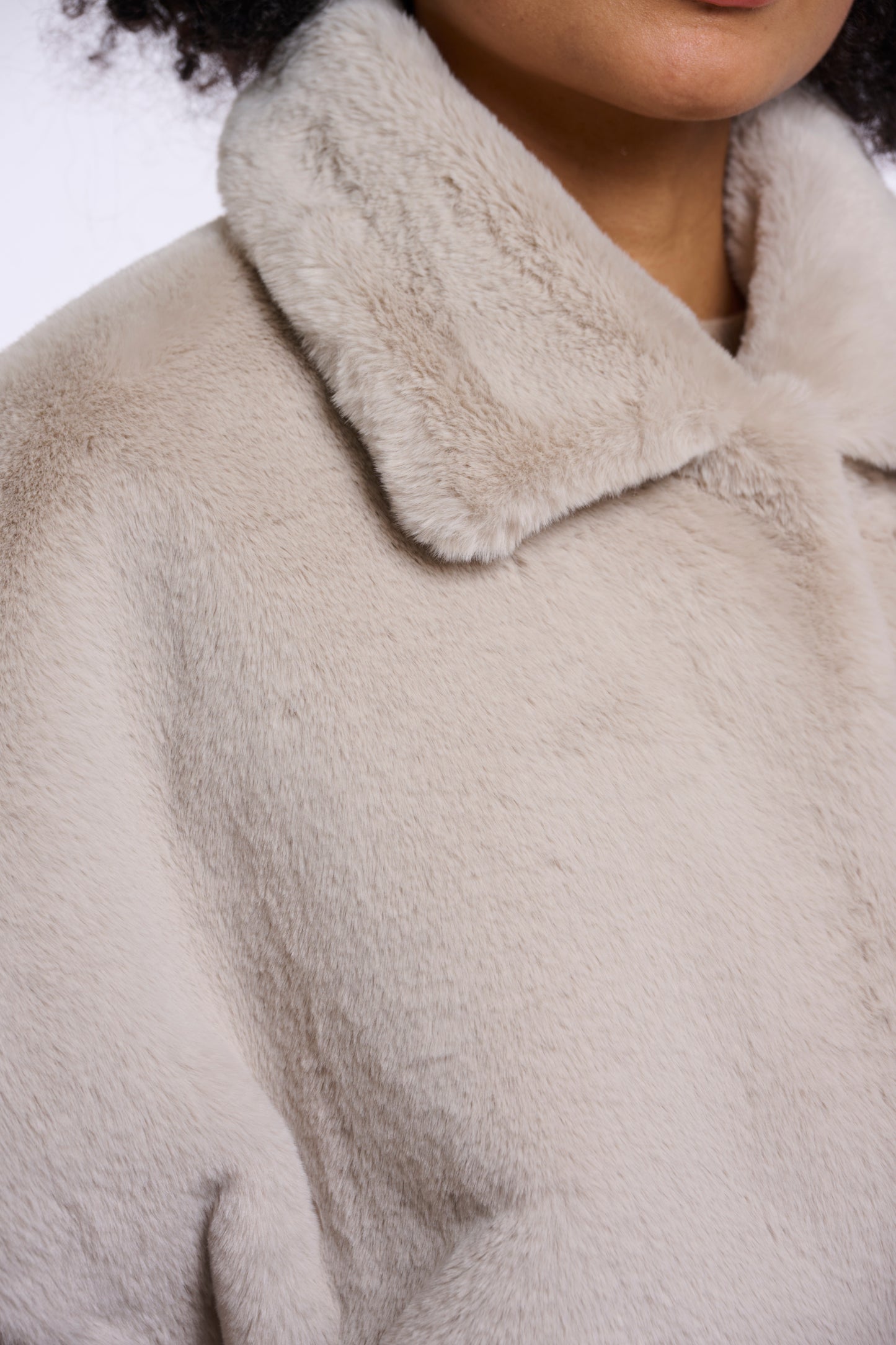Close-up of a person wearing a soft, plush faux-fur or shearling-style coat in a light warm beige tone, showing the collar and textured fabric.