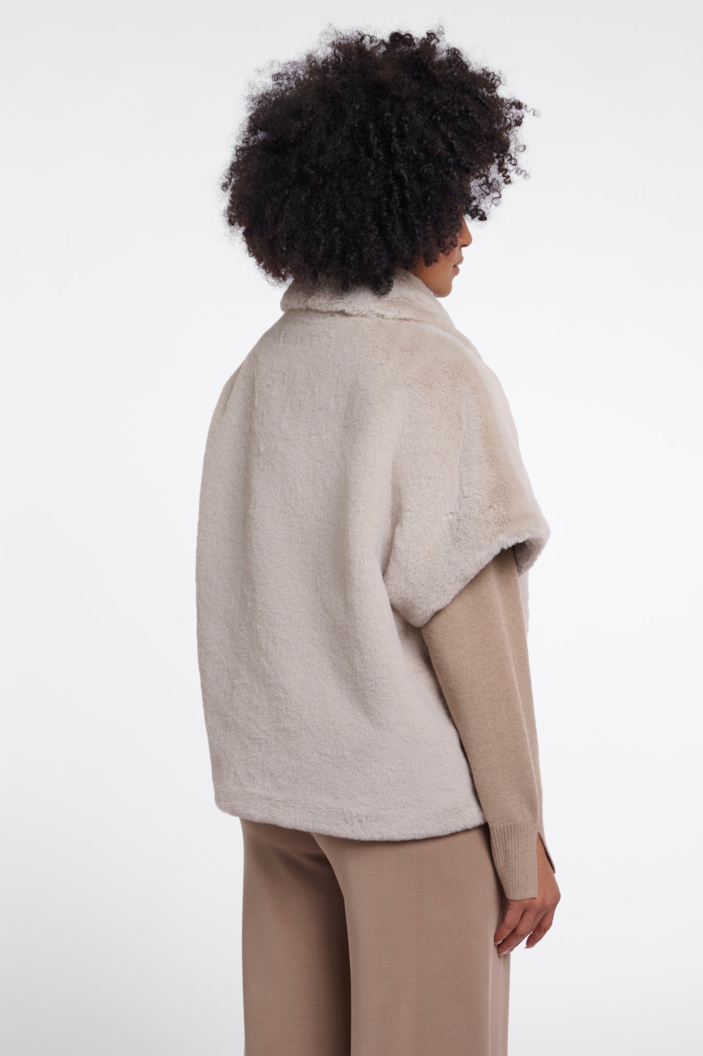 A model shown from the back wearing a light beige fuzzy cropped cape jacket over a tan long-sleeve top and beige trousers, against a plain white studio background.