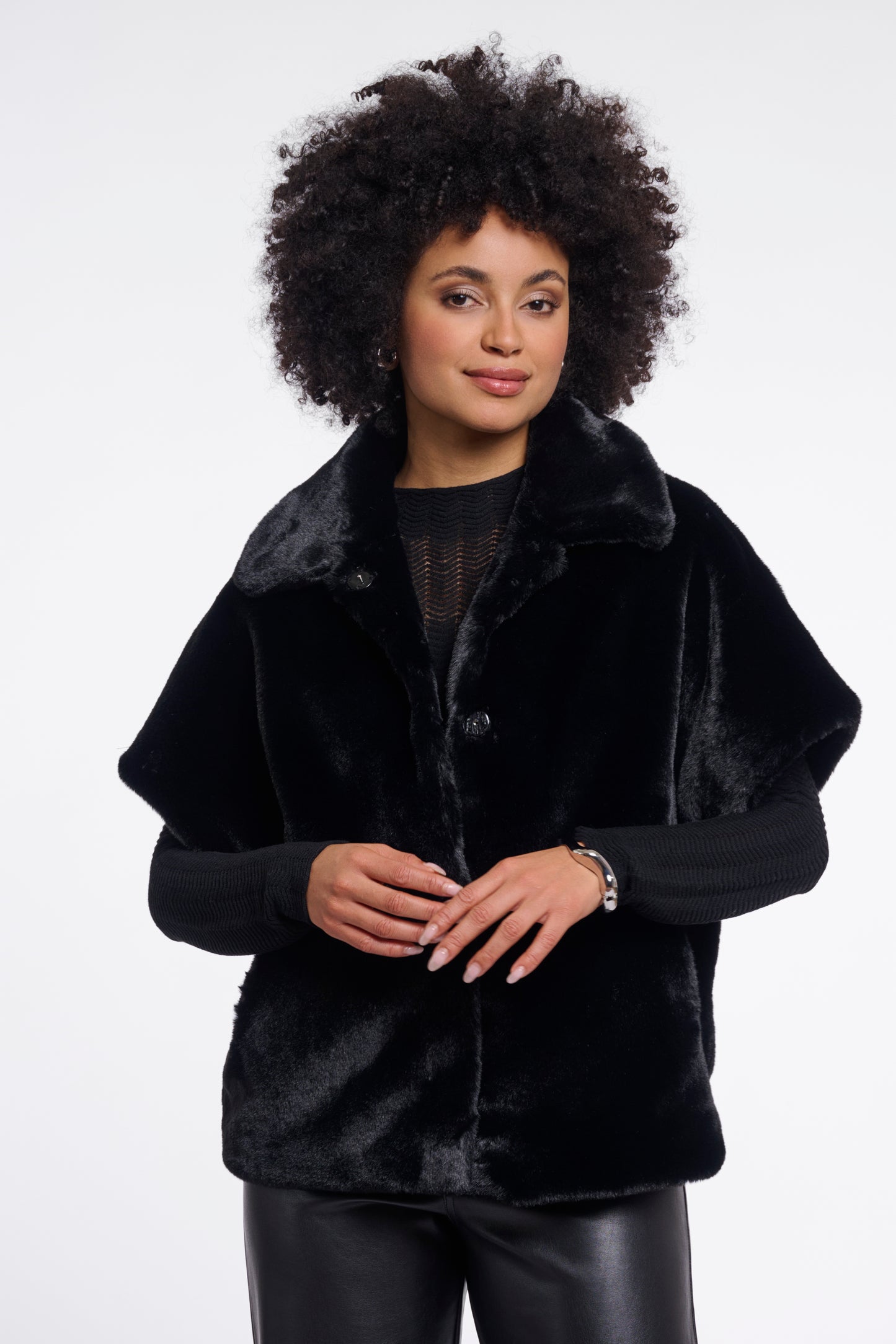 A woman with an afro hairstyle wearing a black plush faux fur cape jacket over a black top and black leather pants, photographed against a plain white studio background.