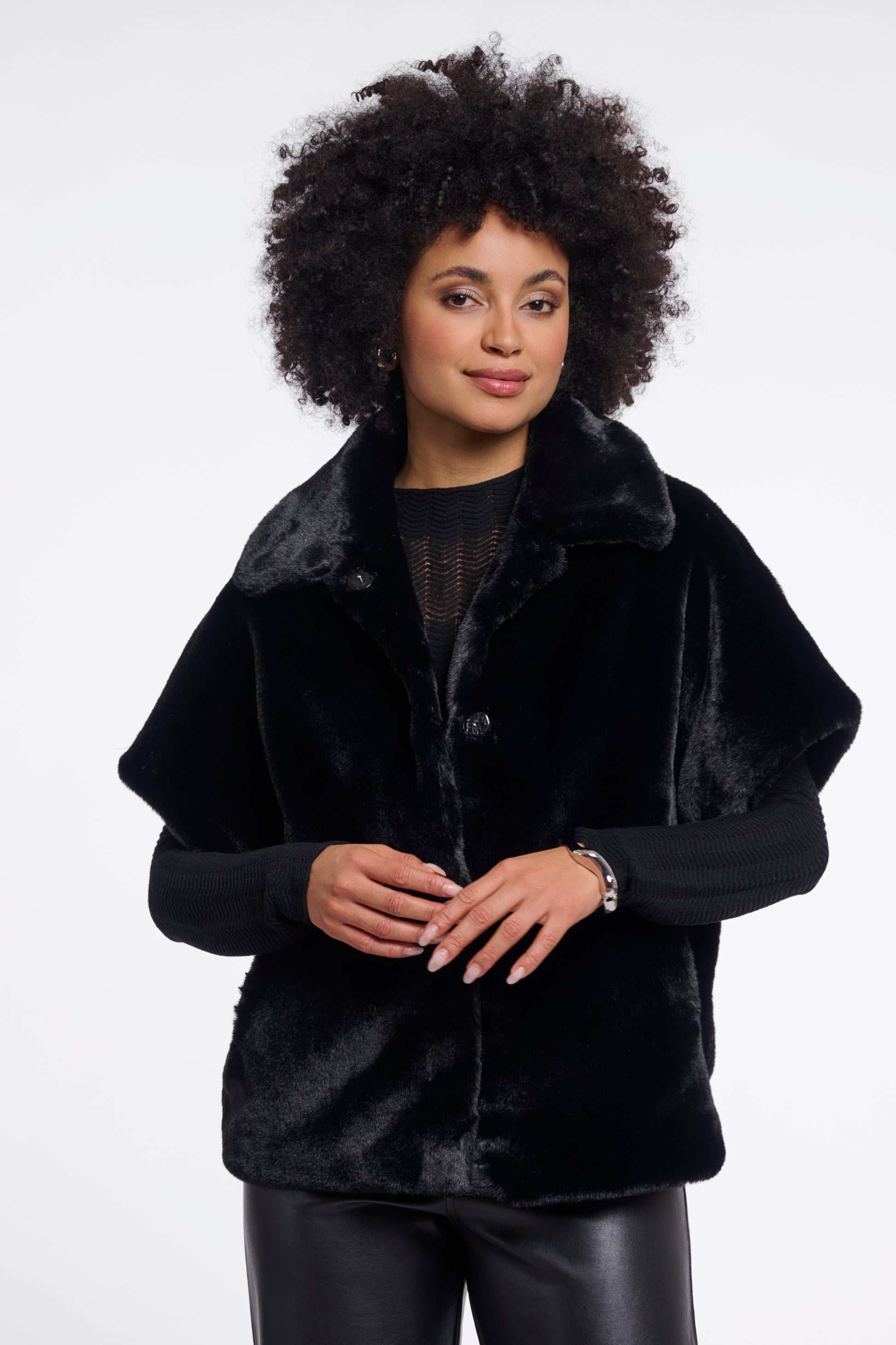 A woman with an afro hairstyle wearing a black plush faux fur cape jacket over a black top and black leather pants, photographed against a plain white studio background.