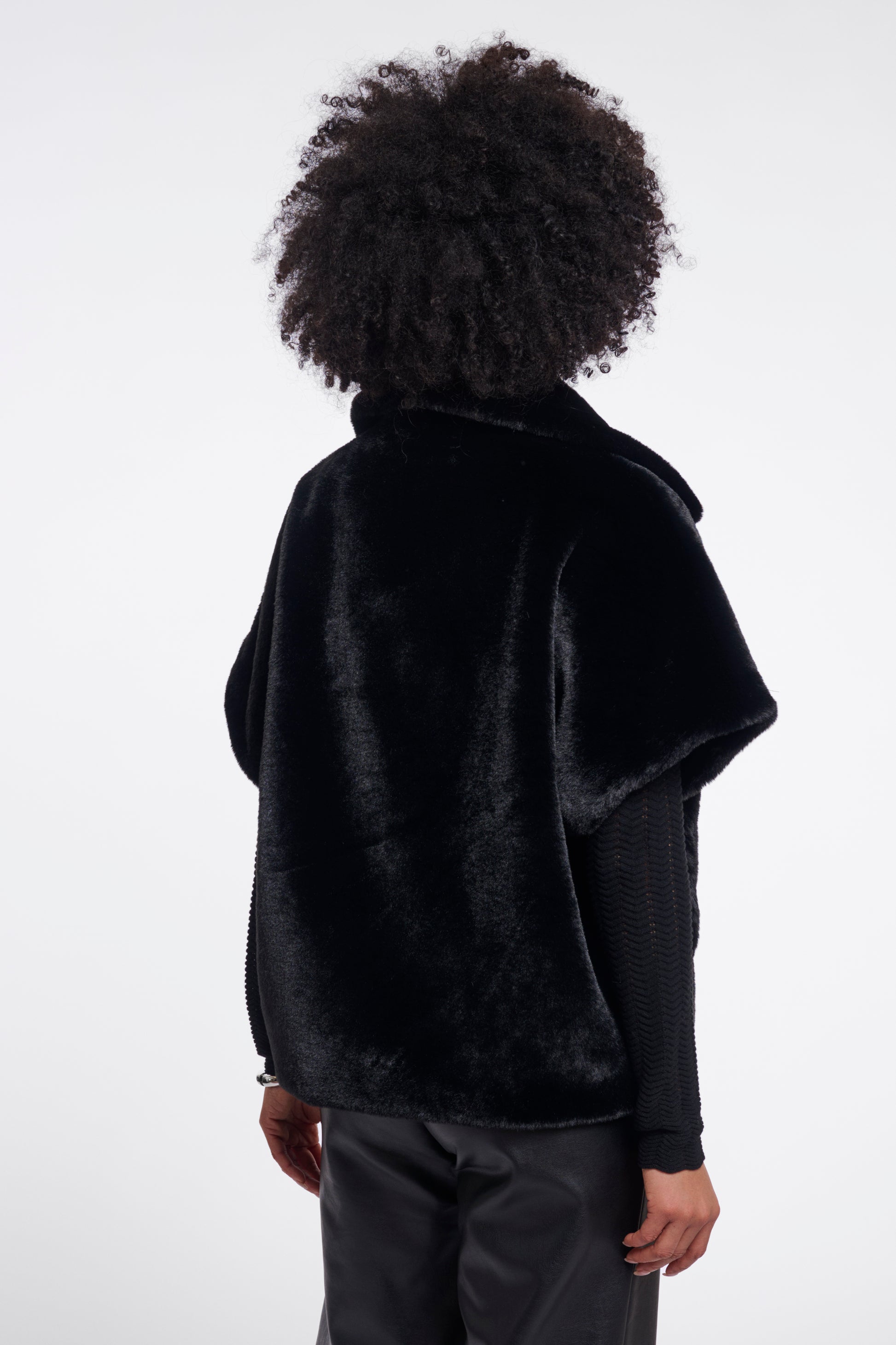 A person shown from the back wearing a black fuzzy cropped jacket or cape over a black long-sleeve top and dark pants, against a plain light gray studio background.