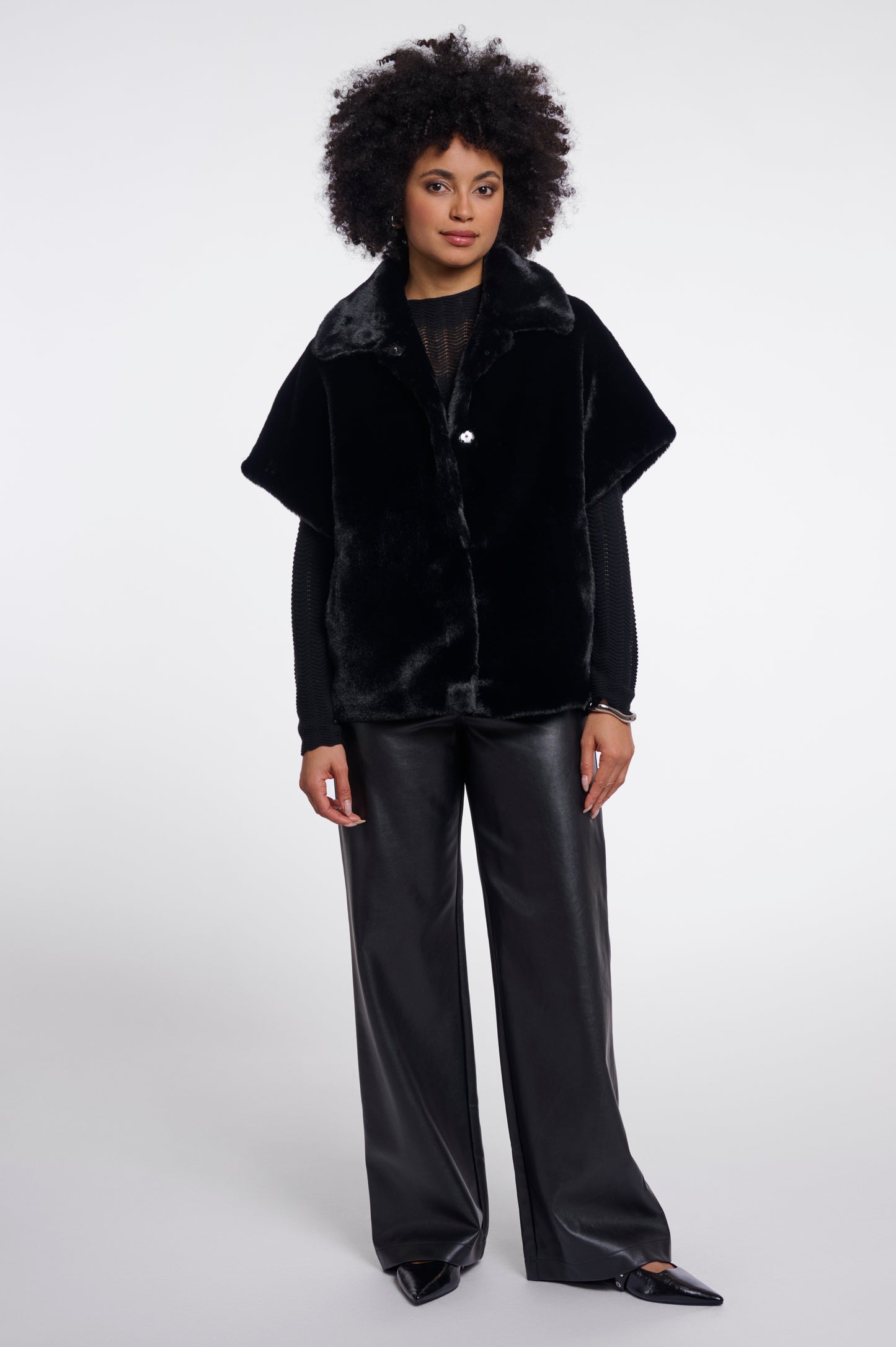 A model with an afro hairstyle wearing a black plush faux-fur cropped jacket with wide sleeves and black wide-leg faux-leather pants, standing against a light gray studio background.
