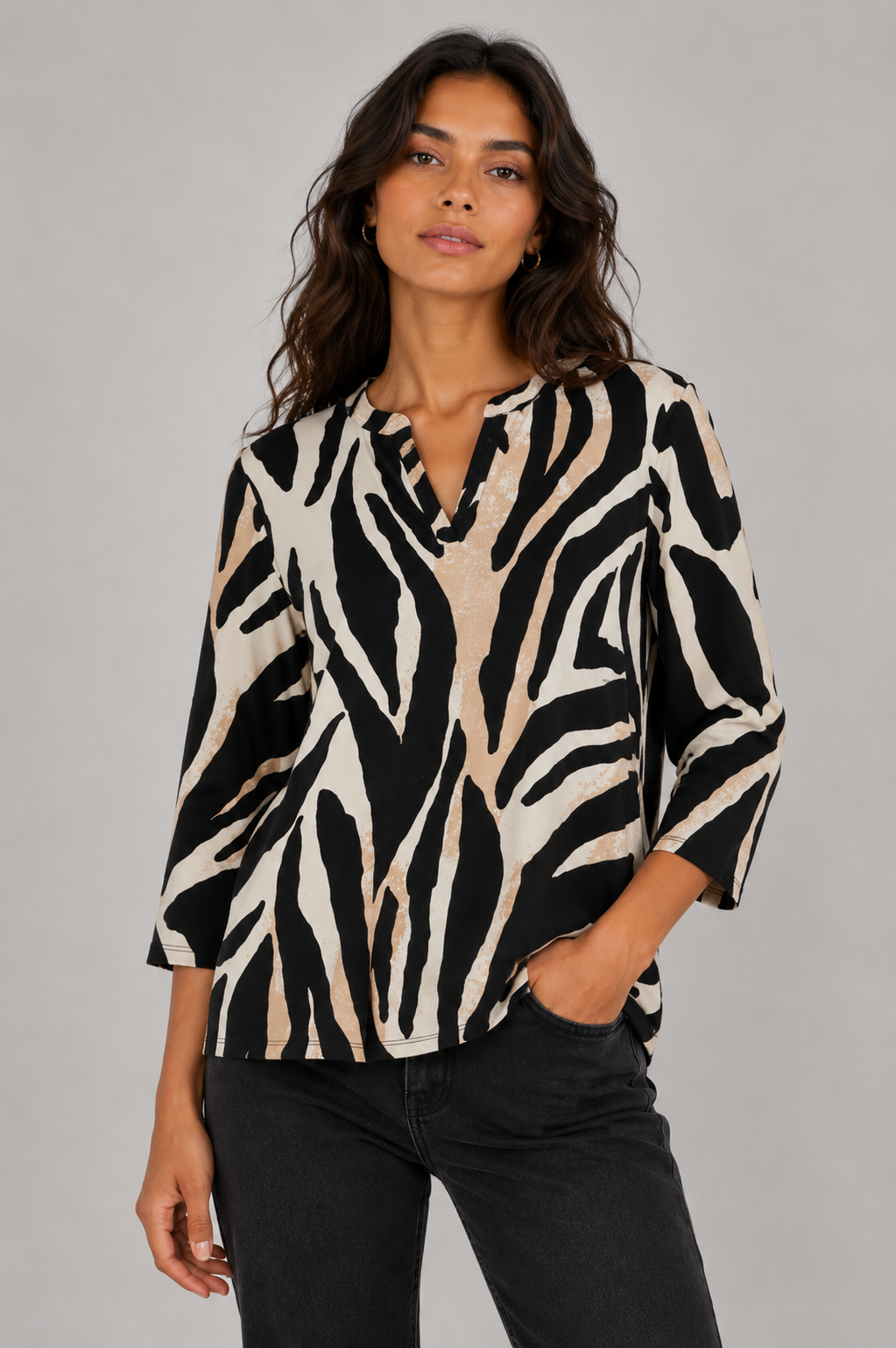 A woman wearing a black and beige zebra-print blouse with three-quarter sleeves and a split neckline, paired with black jeans, against a solid gray studio background.