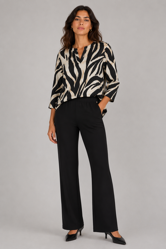 A woman modeling a black and beige zebra-print blouse with three-quarter sleeves, paired with black wide-leg trousers and black heels, against a solid gray studio background.