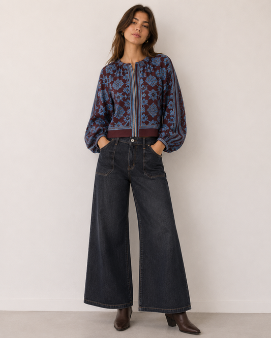 A woman stands against a plain off-white wall wearing a cropped, long-sleeve patterned jacket with blue and burgundy tones, dark wide-leg denim jeans, and brown ankle boots.