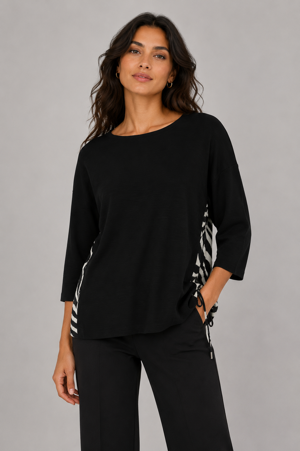 A woman modeling a black three-quarter sleeve top with black and white zebra-patterned side panels and drawstrings, paired with black pants, against a solid gray studio background.