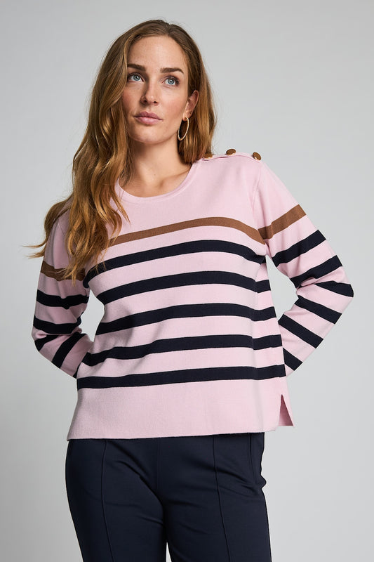 A woman modeling a light pink long-sleeve knit sweater with horizontal navy blue stripes and a thin brown stripe across the chest, paired with dark navy pants, against a neutral gray studio background.
