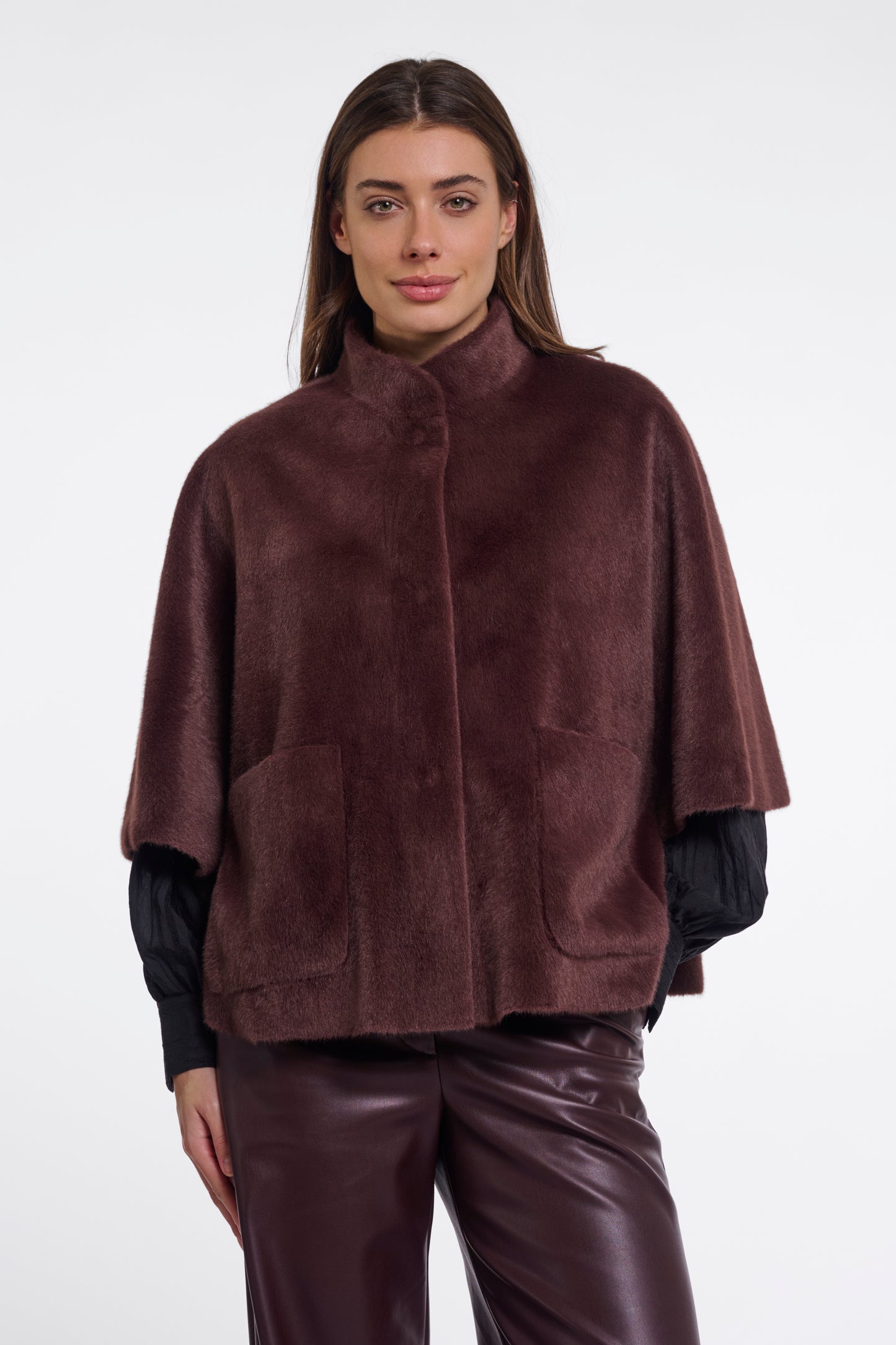 A woman modeling a dark brown faux fur cape jacket with a stand-up collar and front pockets, worn over a black long-sleeve top and paired with matching dark brown faux leather pants, against a plain white studio background.