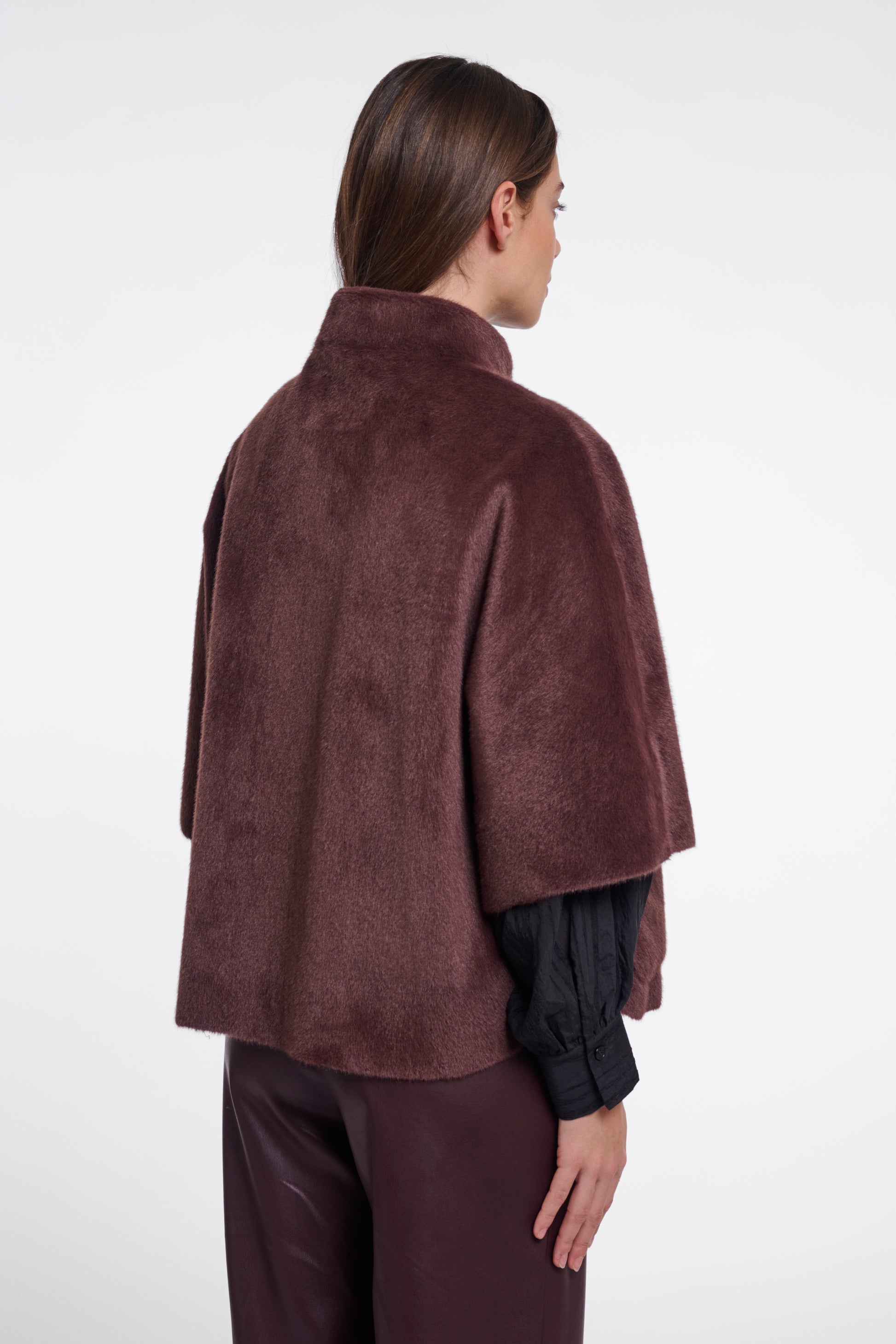 A model shown from the back wearing a dark brown plush faux-fur cape jacket over a black long-sleeve top and matching dark trousers, against a plain white studio background.