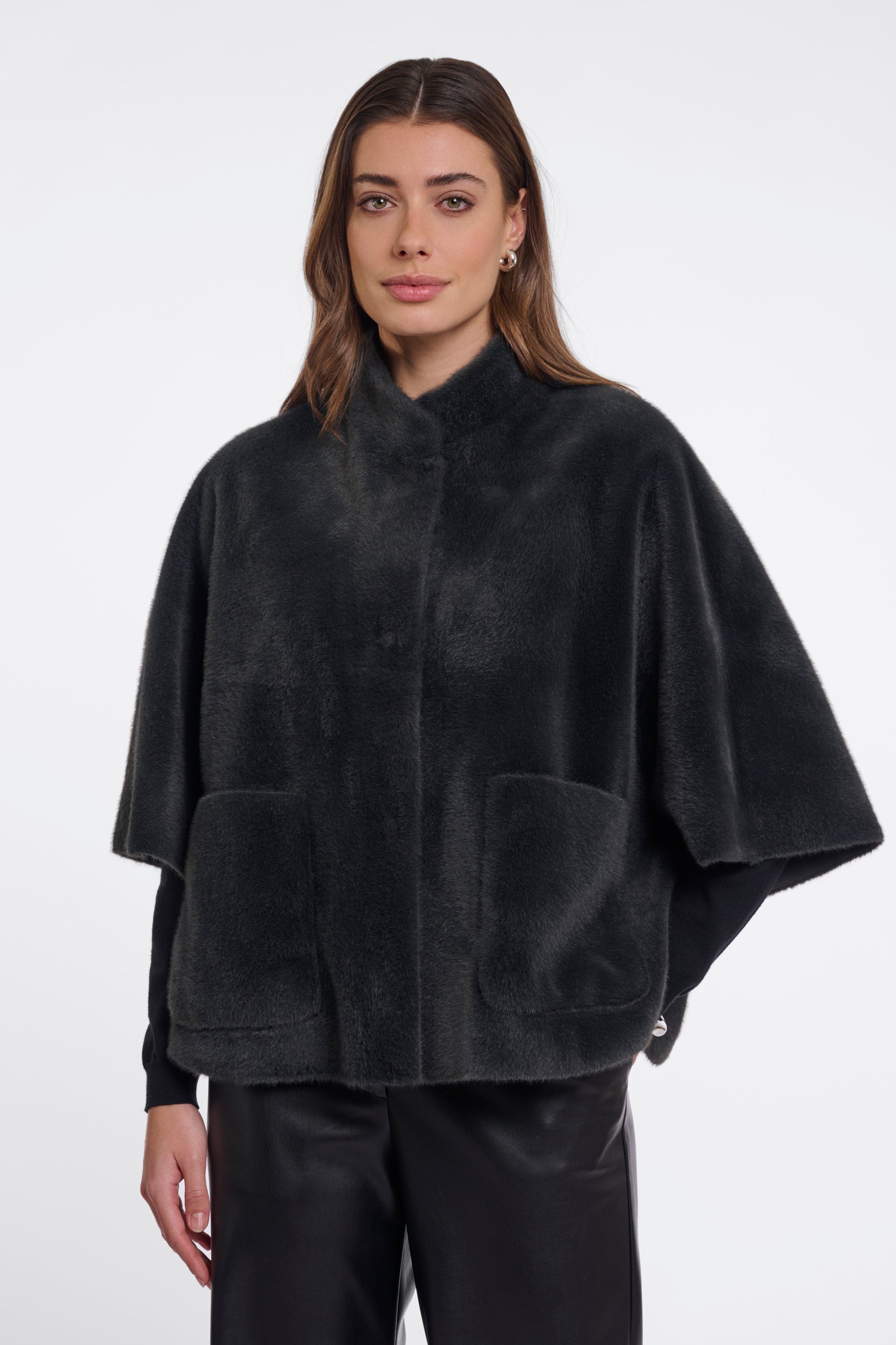 A woman modeling a black plush faux-fur cape jacket with wide draped sleeves, a stand collar, and two large front patch pockets, photographed against a clean light gray studio background.