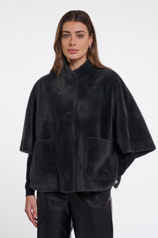 A woman modeling a black plush faux-fur cape jacket with wide draped sleeves, a stand collar, and two large front patch pockets, photographed against a clean light gray studio background.