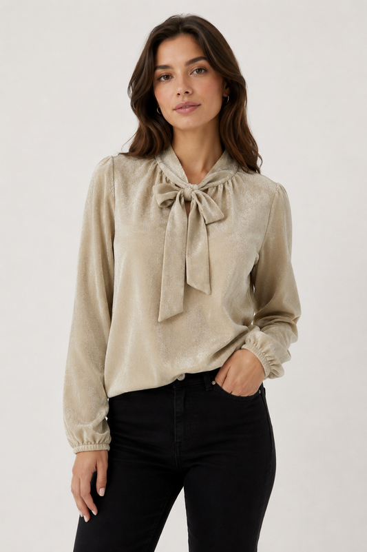 A woman wearing a beige long-sleeve blouse with a large bow tie neckline and black pants, standing against a plain light gray studio background.