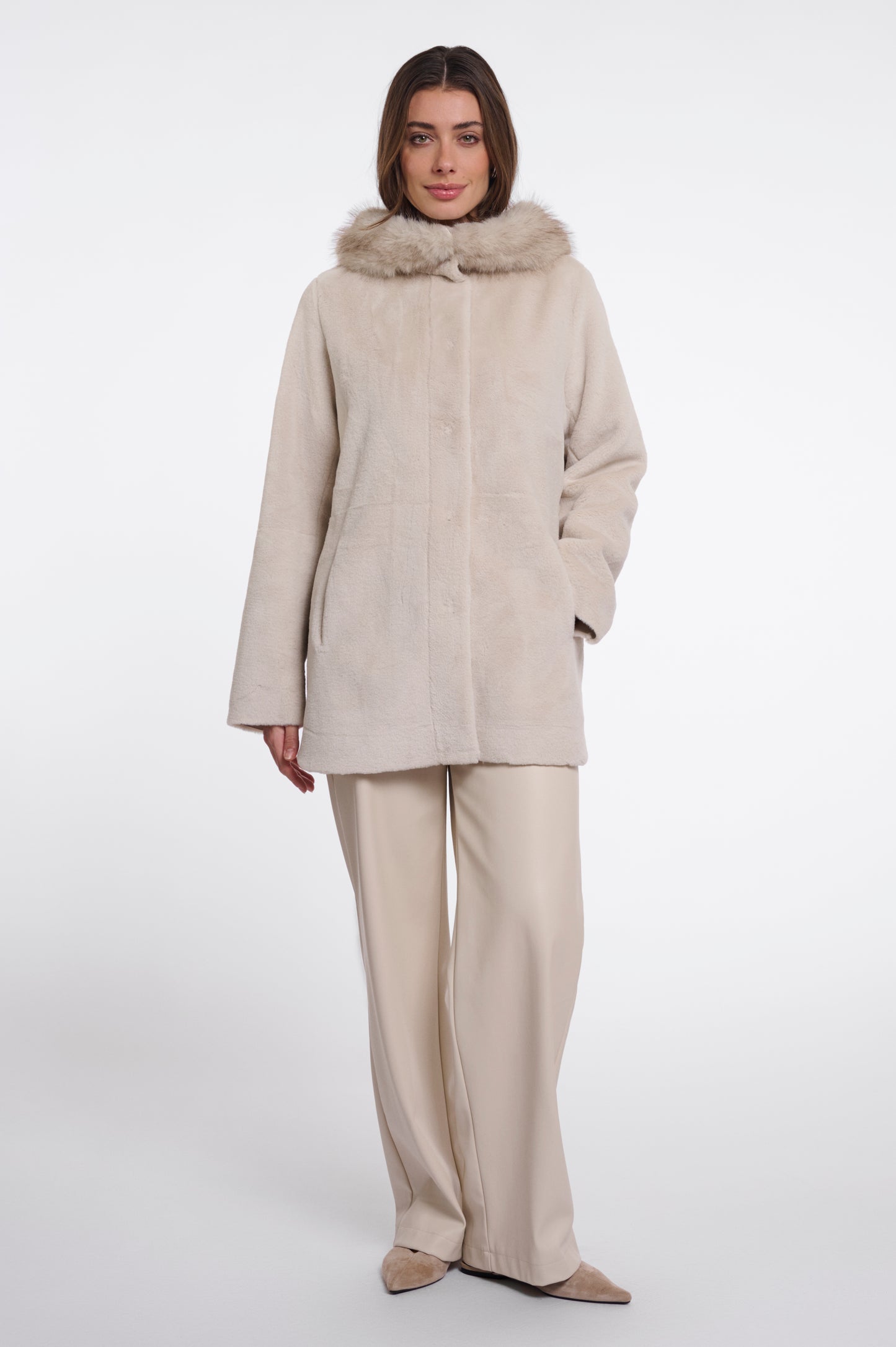 A woman modeling a light beige faux fur coat with a matching fur-trimmed collar and wide-leg beige trousers against a plain light gray studio background.