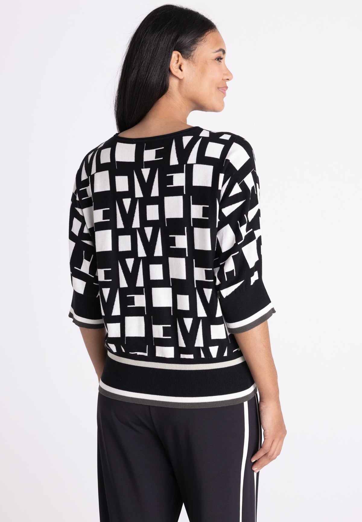 A woman shown from the back wearing a black and white geometric patterned sweater with three-quarter sleeves and black pants with white side stripes, against a plain light gray studio background.
