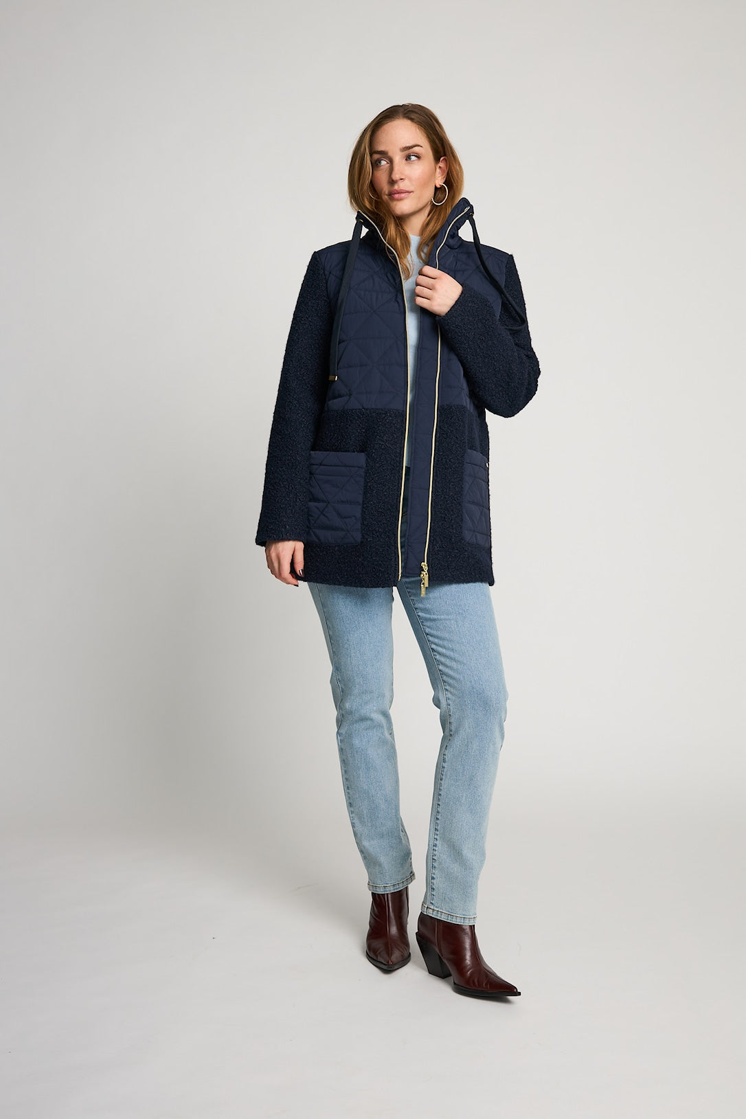 A woman modeling a navy blue patchwork quilted jacket with a gold zipper, paired with light blue jeans and dark brown ankle boots, against a light gray studio background.