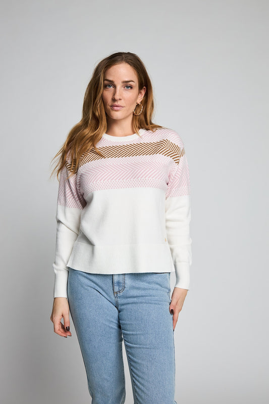 A woman modeling a color-block knit sweater with a white body, light pink upper sleeves, and a beige chevron pattern across the chest, paired with light blue jeans against a plain gray studio background.