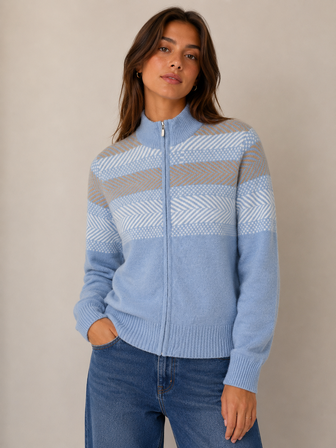 A woman wearing a light blue zip-up knit sweater with beige and white patterned horizontal stripes, paired with blue jeans, standing against a neutral beige background.