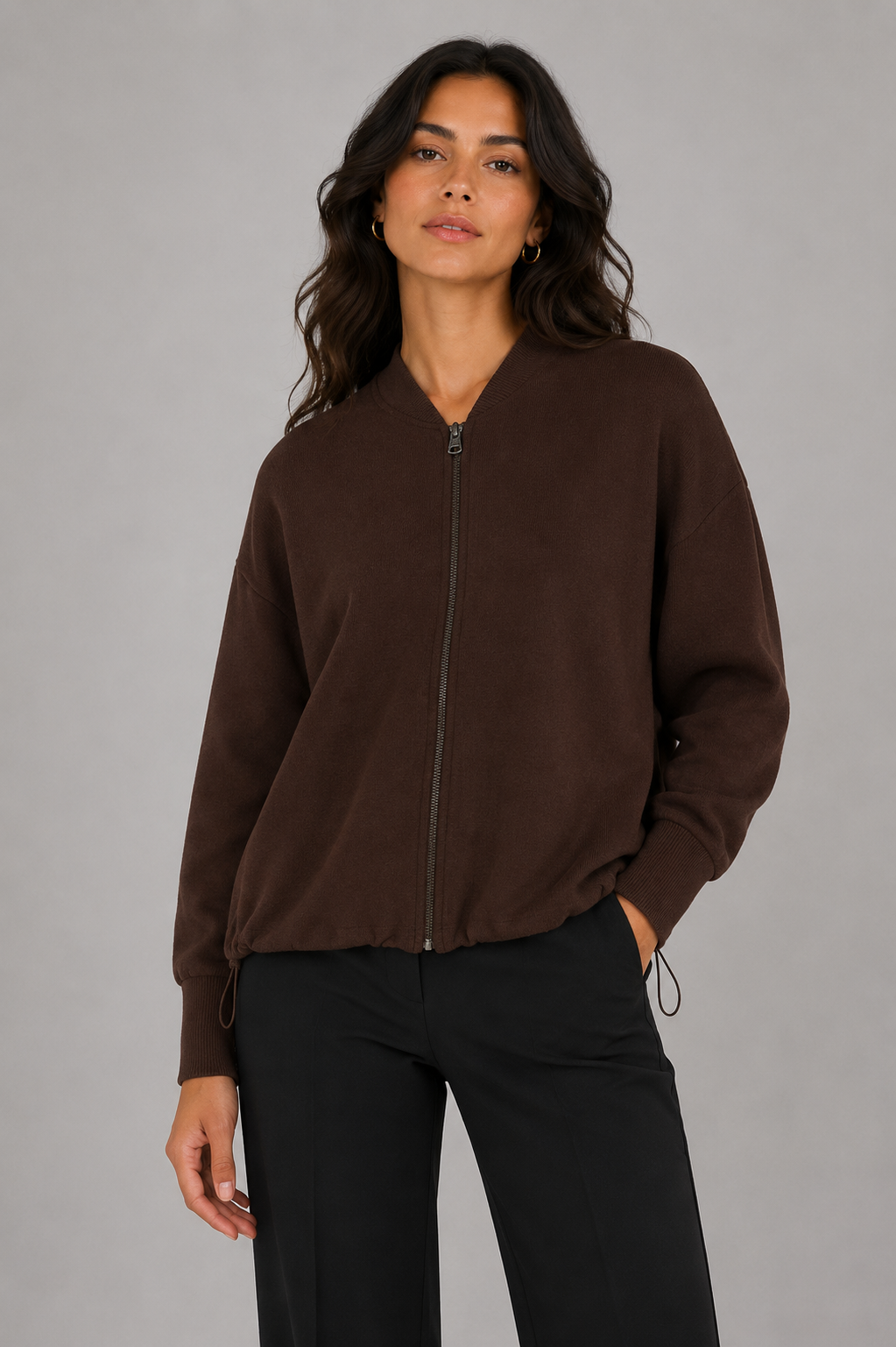 A woman wearing a dark brown zip-up knit sweater with side drawstrings and black pants, standing against a solid gray studio background.