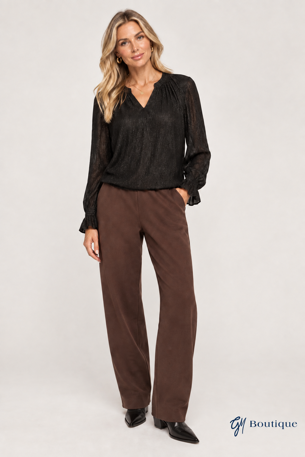 AI-generated styling mockup, not product photography: black Mos Mosh Ximena Glam blouse styled with brown MMARDIE Barrel Sweatpants, based on the store's product images. Trouser details may vary. Small GM Boutique logo at bottom right.