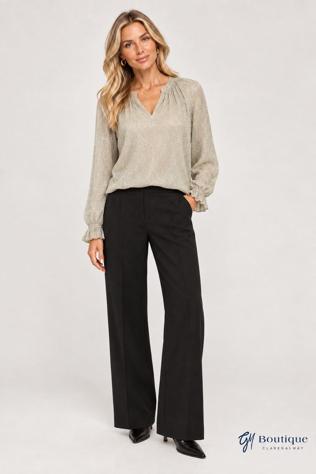 AI-generated styling mockup, same light-skinned blonde model wearing the gold Mos Mosh Ximena Glam blouse with black Oui wide-leg tailored trousers; fine metallic fibre surface and small navy GM Boutique logo.